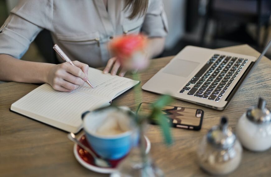 A close up on a desk with a laptop, a cup of coffee, and a journal that someone is writing in.