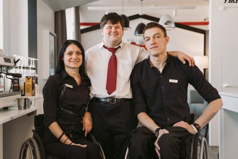 Two people using wheelchairs wearing black work uniforms pose with a man with down syndrome between them who is dressed in work attire. All are smiling for the camera.