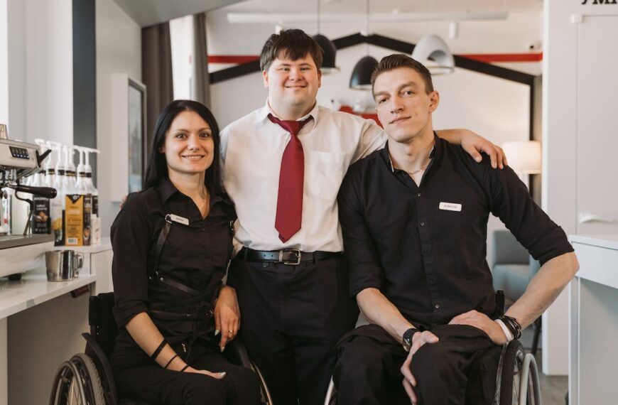 Two people using wheelchairs wearing black work uniforms pose with a man with down syndrome between them who is dressed in work attire. All are smiling for the camera.