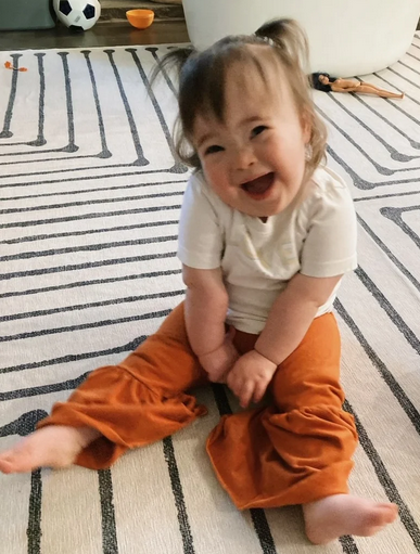 A baby girl smiles up at the camera. She's wearing orange pants and has her hair in small pigtails.