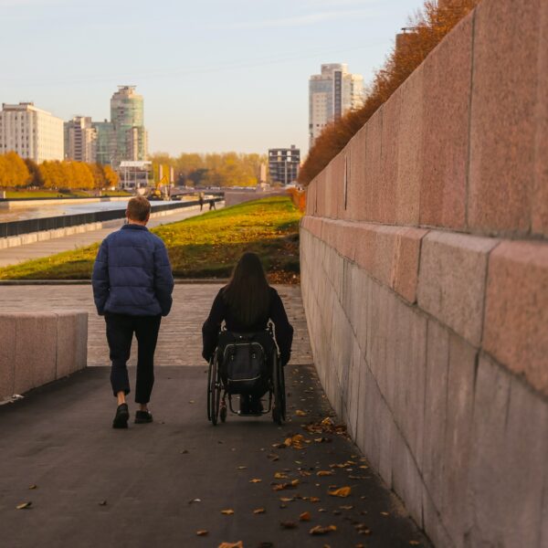 A man and a woman in a wheelchair go down a ramp to stroll by the river near a bright city