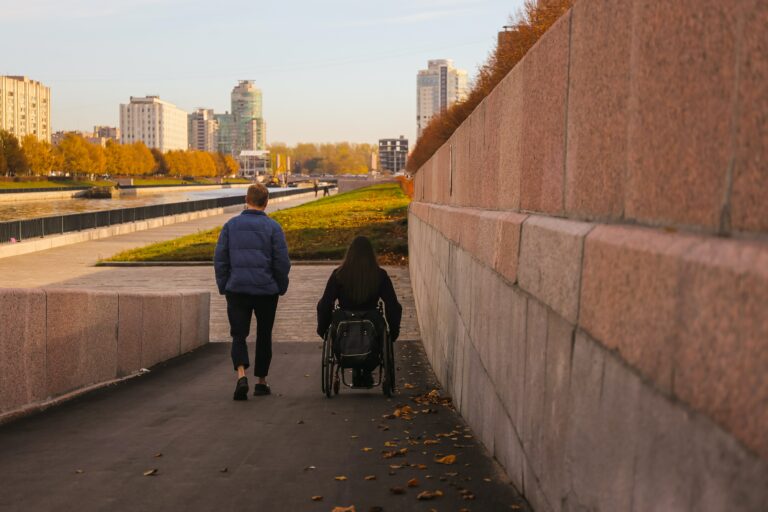 A man and a woman in a wheelchair go down a ramp to stroll by the river near a bright city