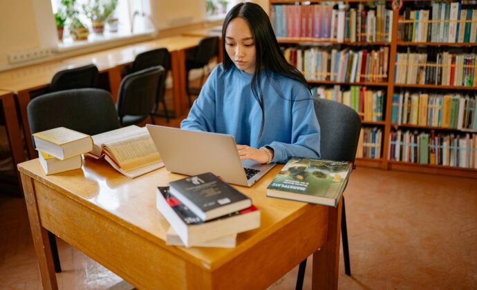A woman sits in a library at a table with a computer and multiple books