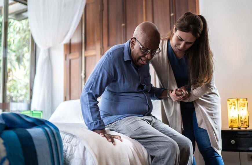 A young woman in scrubs assists an elderly man in standing up from his bed.