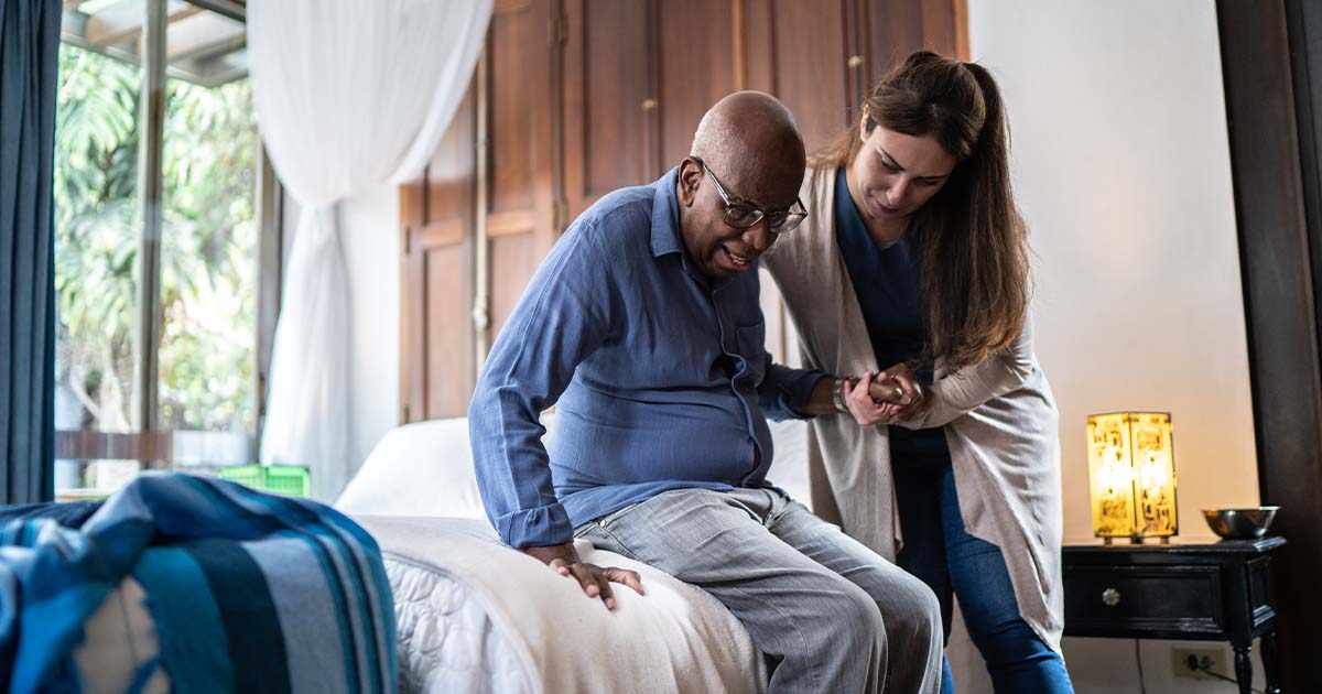 A young woman in scrubs assists an elderly man in standing up from his bed.