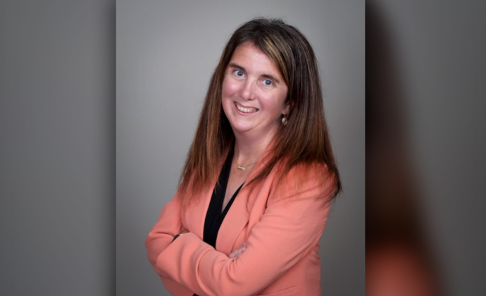 A head shot of Lynn Pascoa - Brown haired, blue eyed woman smiling with crossed arms wearing a salmon jacket