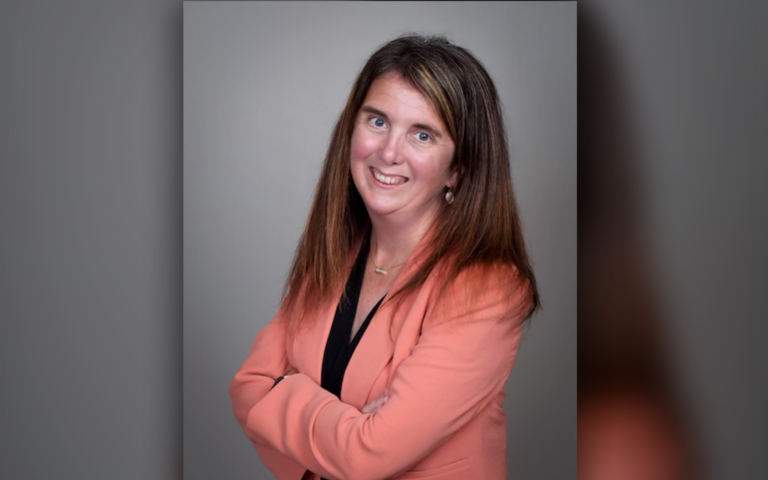 A head shot of Lynn Pascoa - Brown haired, blue eyed woman smiling with crossed arms wearing a salmon jacket