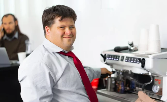 A man with down syndrome wearing a read tie makes coffee while smiling at the camera