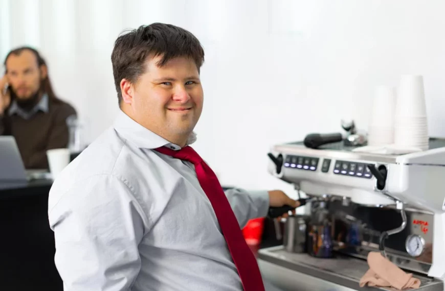 A man with down syndrome wearing a read tie makes coffee while smiling at the camera