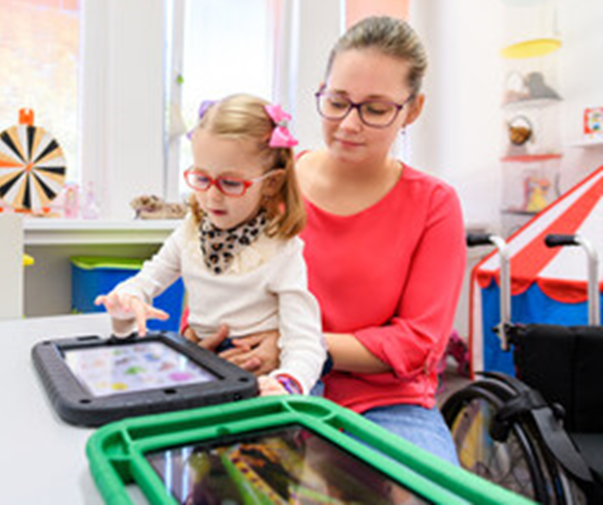 A little girl operates a tablet displaying communication pictures  while sitting on her mother's lap