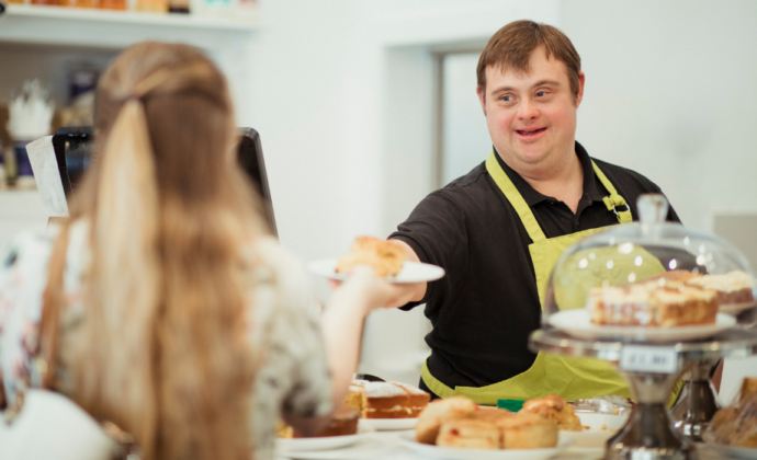 A man with down syndrome wearing a yellow apron hands a plate holding a pastry to a blonde woman across a counter