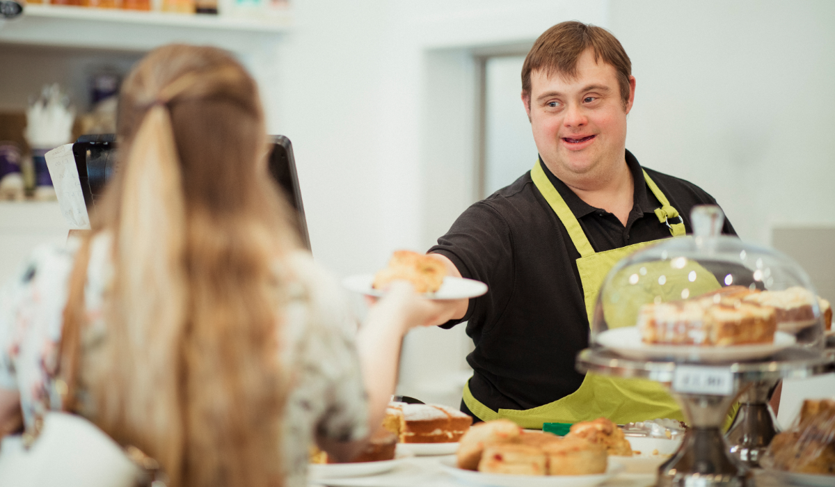 A man with down syndrome wearing a yellow apron hands a plate holding a pastry to a blonde woman across a counter