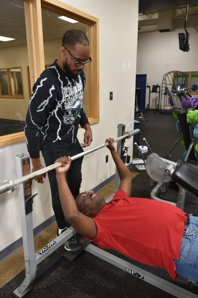 A man lying on a bench press lifts weights while a man standing behind the bench spots him