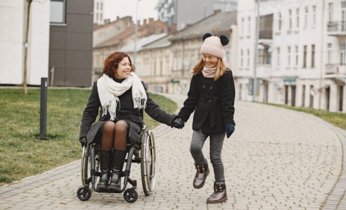 A woman using a wheelchair is bundled up in a coat and smiling at her twelve year old daughter who is wearing a knitted cap while laughing