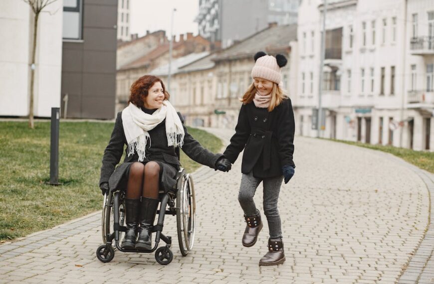 A woman using a wheelchair is bundled up in a coat and smiling at her twelve year old daughter who is wearing a knitted cap while laughing