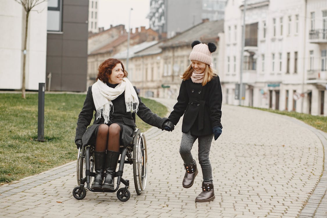 A woman using a wheelchair is bundled up in a coat and smiling at her twelve year old daughter who is wearing a knitted cap while laughing