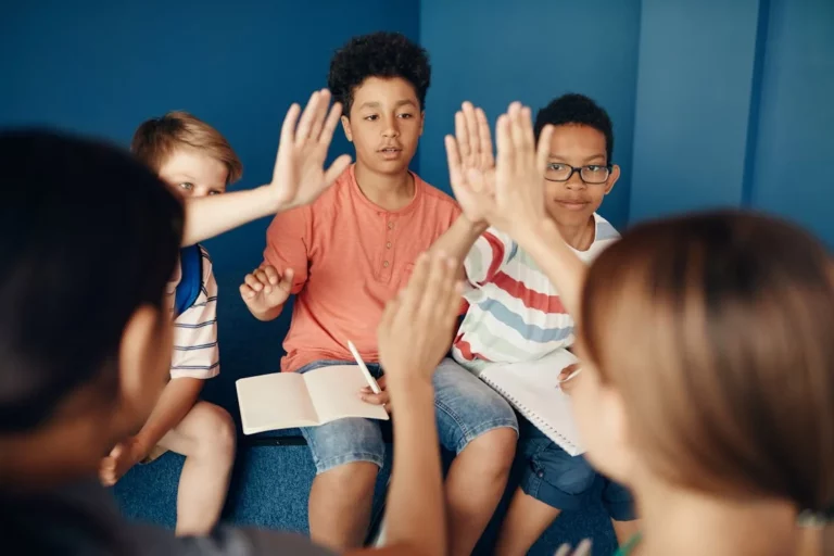 Three children with notebooks in their laps high five two teachers sitting in front of them