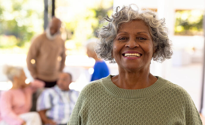 A woman with gray curly hair offers the camera a big white smile in front of a group of seniors.
