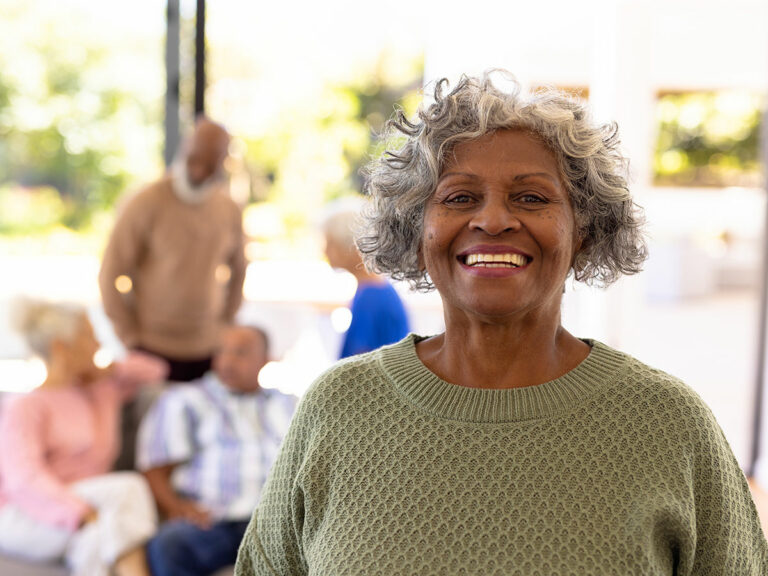 A woman with gray curly hair offers the camera a big white smile in front of a group of seniors.