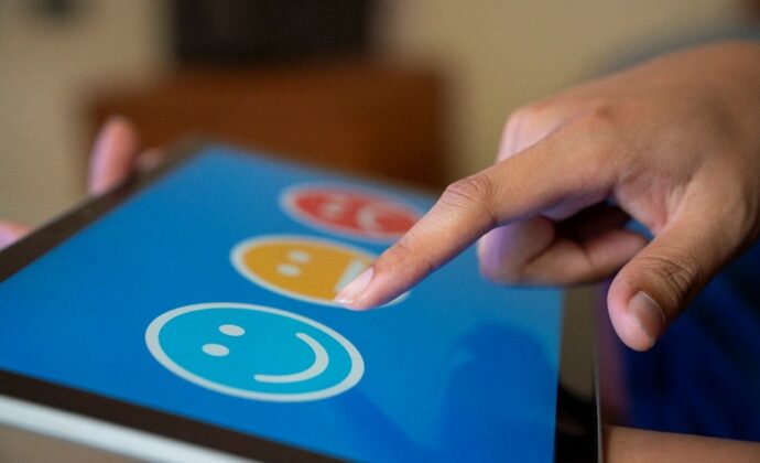 closeup of an ipad screen displaying a happy, neutral, and sad emoji. A finger is selecting the happy emoji.