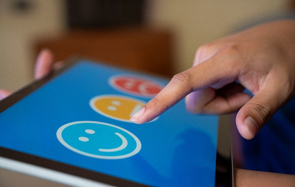 closeup of an ipad screen displaying a happy, neutral, and sad emoji. A finger is selecting the happy emoji.