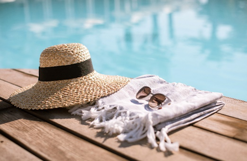 A sun hat, sunglasses, and a pool towel sit on a wood deck next to blue pool waters.