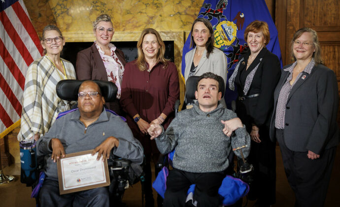 Oscar Drummond, a power chair user, sits in the front row displaying his award with Robert Zoytnia by his side also in a power chair. Council members stand behind them. They are in a stately room with marbled walls and flags.