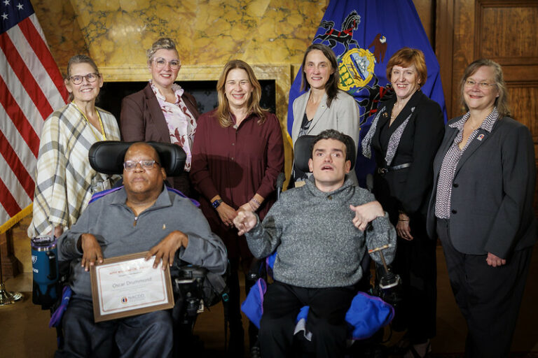 Oscar Drummond, a power chair user, sits in the front row displaying his award with Robert Zoytnia by his side also in a power chair. Council members stand behind them. They are in a stately room with marbled walls and flags.