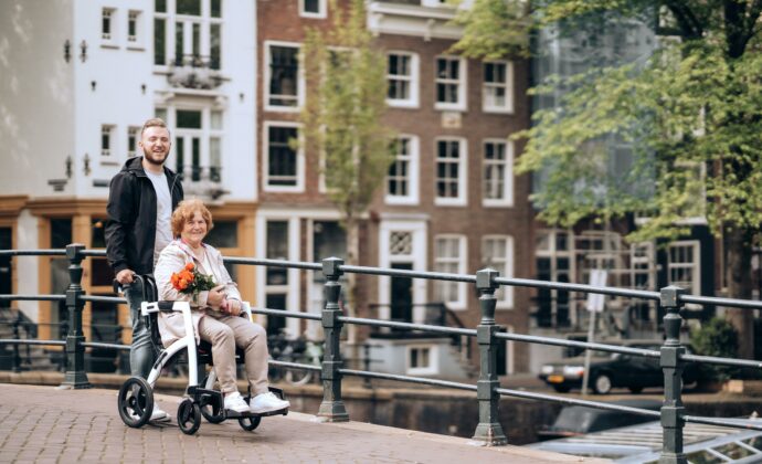 A young man pushes an elderly woman in a wheelchair over a bridge. They are both smiling.