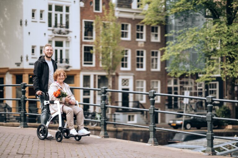 A young man pushes an elderly woman in a wheelchair over a bridge. They are both smiling.