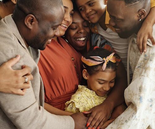 A family embraces in a group hug while smiling. There are two children in the middle of five adults hugging them.