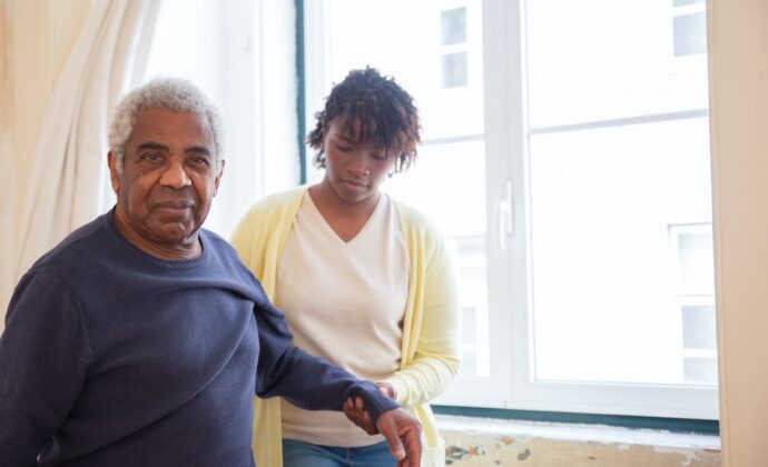 A young woman assists an elderly man in walking by holding his arm to stabilize him. They are standing next to a window.
