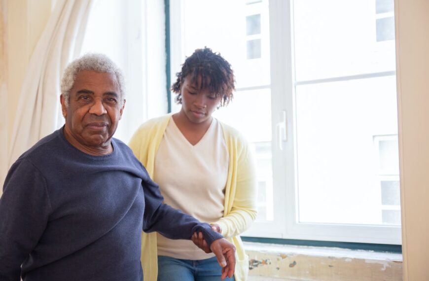 A young woman assists an elderly man in walking by holding his arm to stabilize him. They are standing next to a window.