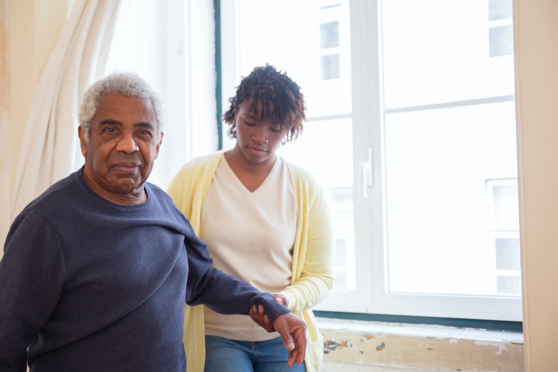A young woman assists an elderly man in walking by holding his arm to stabilize him. They are standing next to a window.