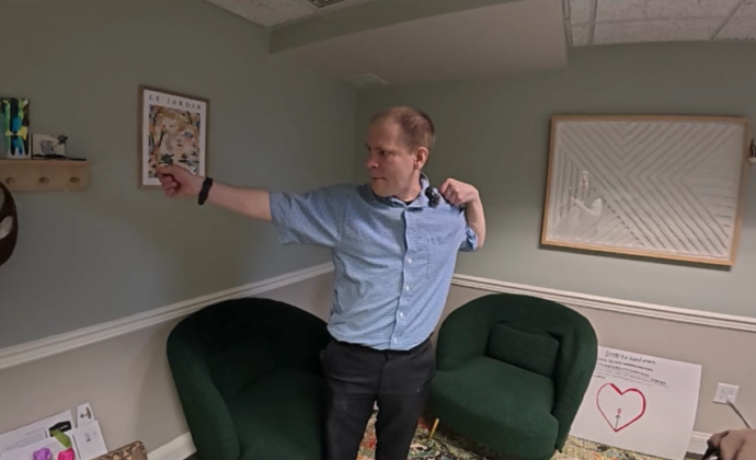 A man in a blue button down shirt stands in a waiting room doing the tai chi pose "Bow and Arrow"