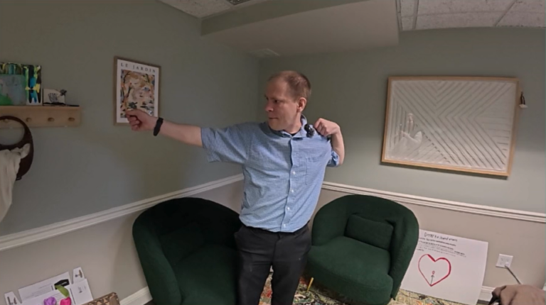A man in a blue button down shirt stands in a waiting room doing the tai chi pose "Bow and Arrow"