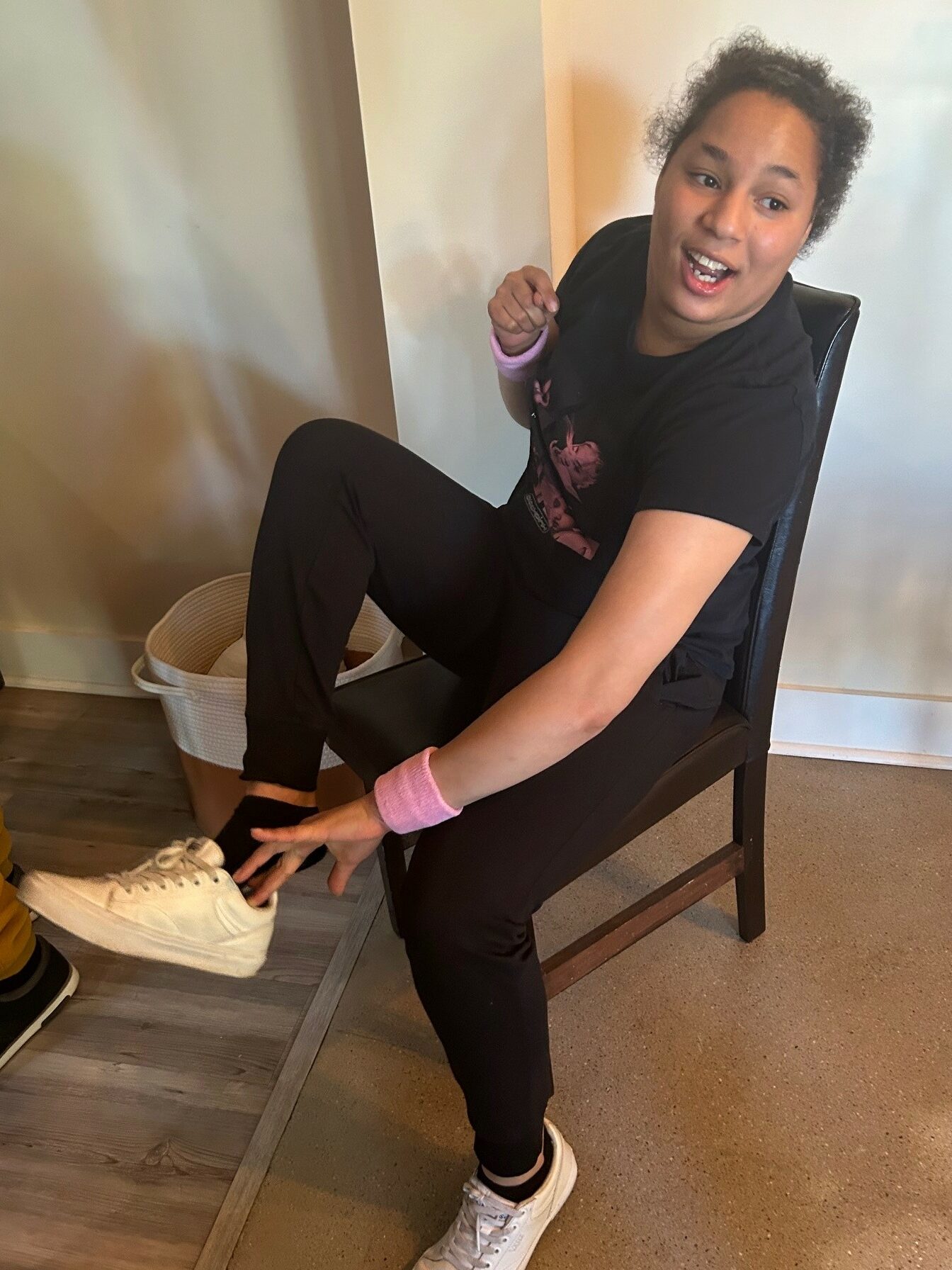 Alexis J - a young woman with black curly hair wearing black athletic wear - sit to put on her white sneakers while talking and smiling to someone out of frame.