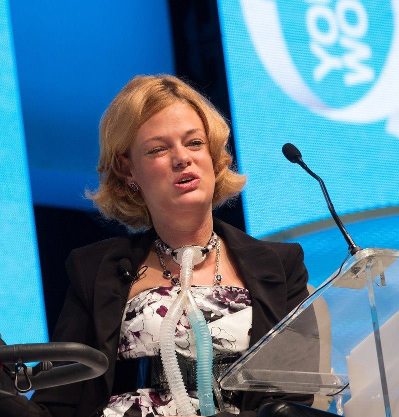 a headshot of Dr. Josie Badger delivering a professional speech on stage. She is woman with blonde hair and brown eyes while wearing a floral blouse and black blazer. She is using a power chair and has a tracheostomy tube.