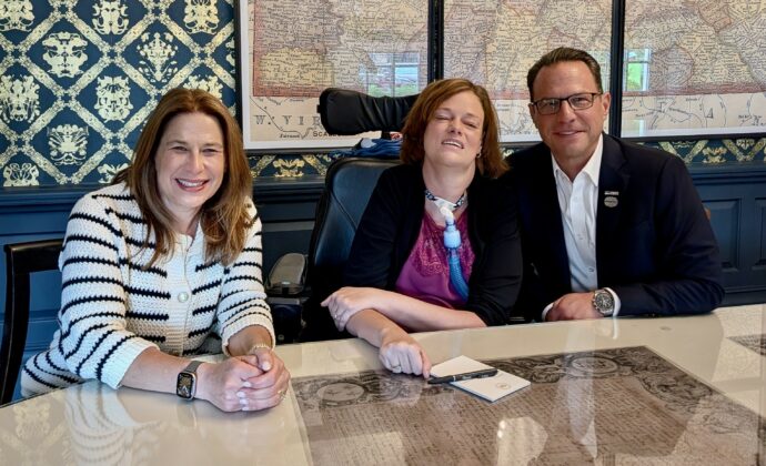 Pennslvania First Lady Lori Shapiro, Dr. Josie Badger, and Governor Josh Shapiro sitting behind a glass table smiling for the camera.