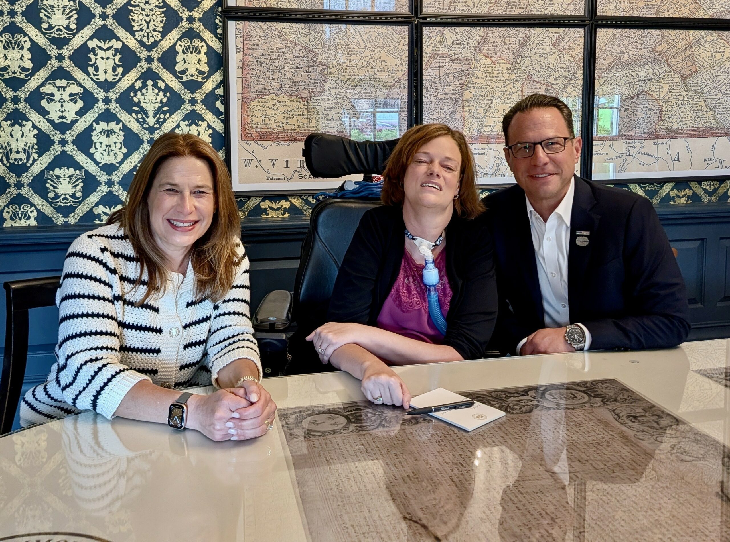 Pennslvania First Lady Lori Shapiro, Dr. Josie Badger, and Governor Josh Shapiro sitting behind a glass table smiling for the camera.